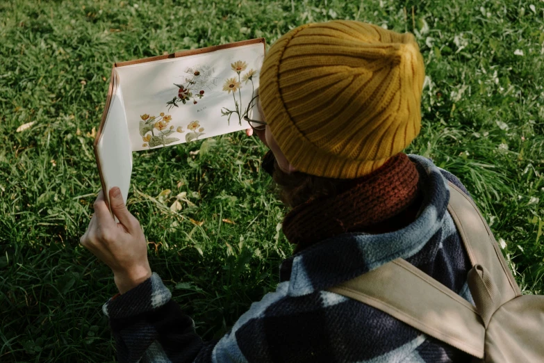 a person sitting in the grass reading a book, a photorealistic painting, inspired by Elsa Beskow, pexels contest winner, visual art, botanical herbarium paper, wearing a yellow hoodie, caracter with brown hat, guide