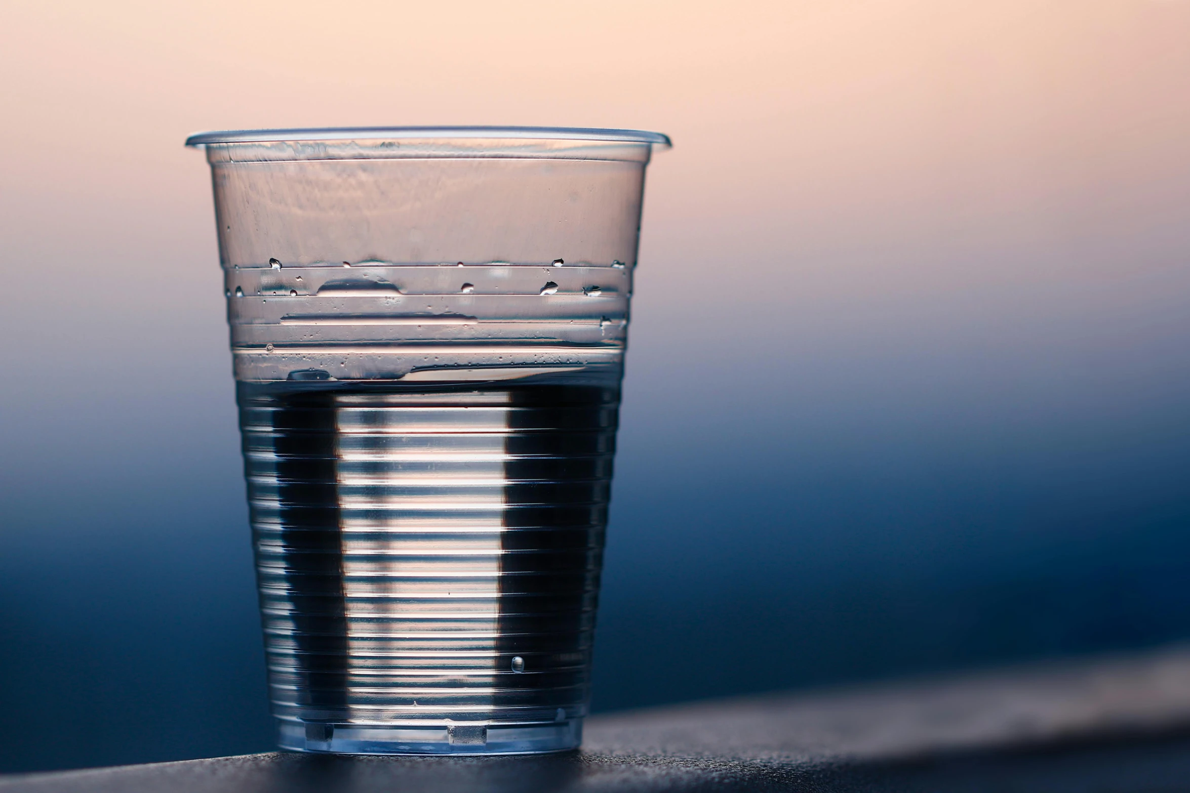 a glass of water sitting on top of a table, unsplash, photorealism, shiny plastic, evening at dusk, paper cup, black water