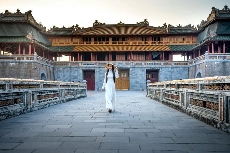 a woman standing on a bridge in front of a building, inspired by Gu An, pexels contest winner, wearing long royal robe, vietnam, square, panoramic centered view of girl