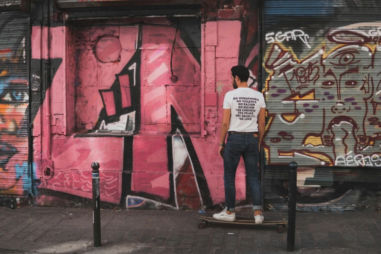 a man standing on a skateboard in front of a pink building, pexels contest winner, graffiti, jeans and t shirt, hidden message, standing with her back to us, young spanish man