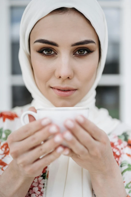 a woman in a hijab holding a cup of coffee, inspired by Maryam Hashemi, pexels contest winner, attractive symmetrical face, tea party, caucasian, white