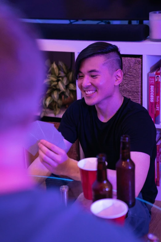 a man sitting at a table talking to another man, by Reuben Tam, drinking beer and laughing, profile image, darren quach, playing cards