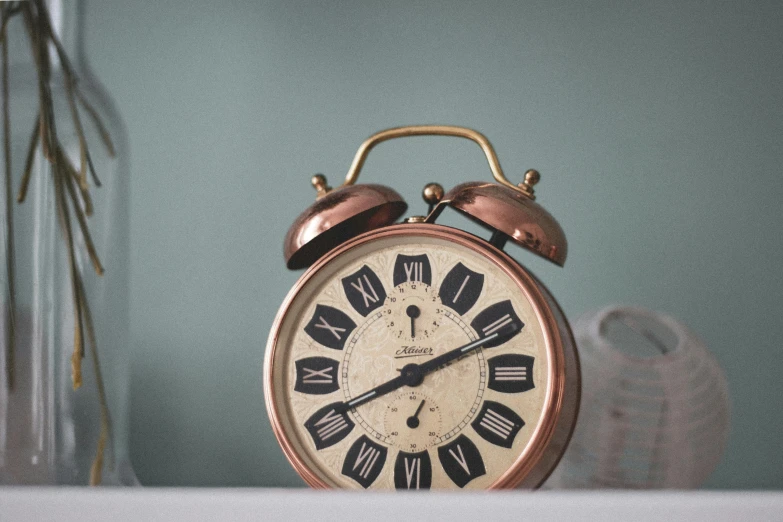 a clock sitting on top of a table next to a vase, inspired by William Nicholson, unsplash, copper and brass, alarm clock, close up front view, vintage style