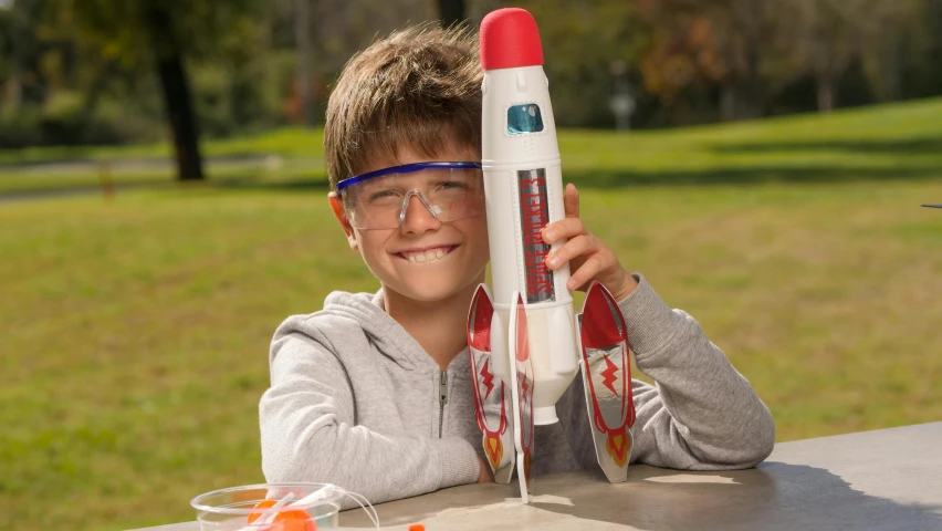 a young boy sitting at a table holding a toy rocket, with navigator shaped glasses, jetfire, full product shot, pictured from the shoulders up