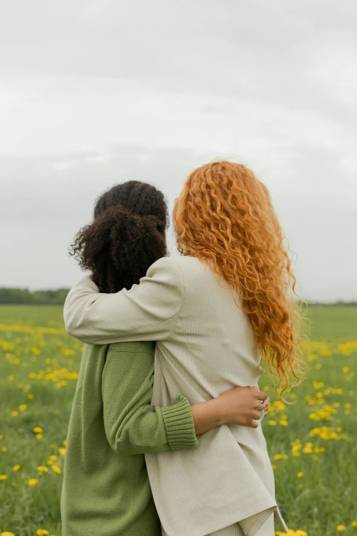 a couple of women standing next to each other in a field, hugging, frizzy hair, varying ethnicities, from the back