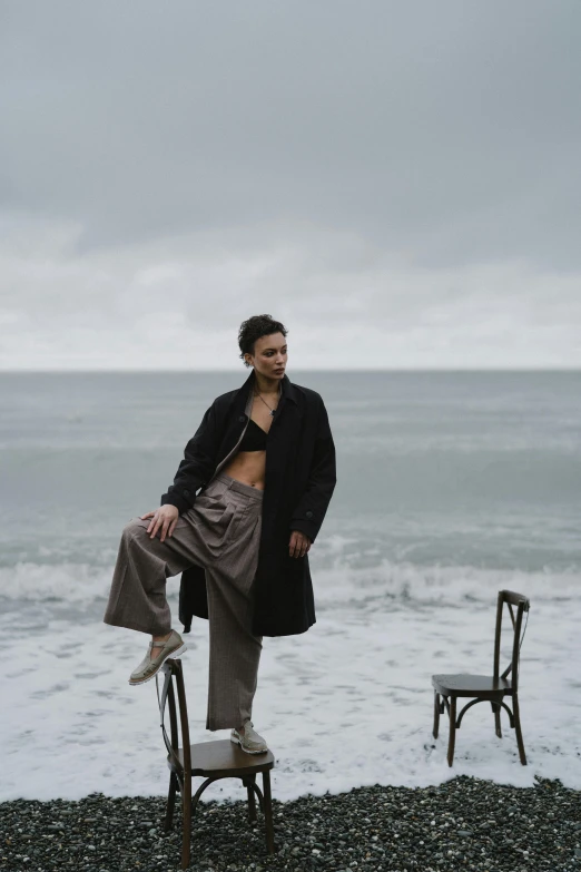 a man standing on top of a chair next to the ocean, a portrait, inspired by Peter Lindbergh, unsplash, woman is in a trenchcoat, infp young woman, black sand, non binary model