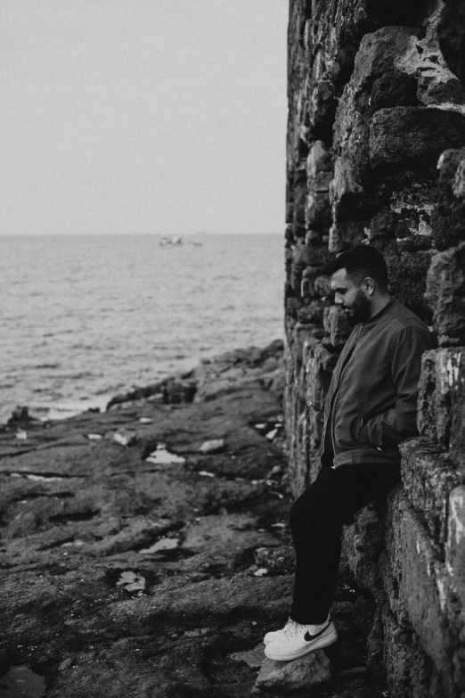 a man sitting on a rock next to the ocean, a black and white photo, profile image, liam, ruins, sad men