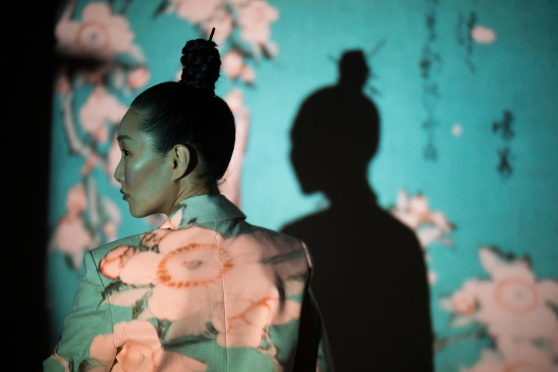 a woman standing in front of a wall with flowers on it, inspired by Otake Chikuha, projection mapping, shadows, with dark hair tied up in a bun, photographed for reuters