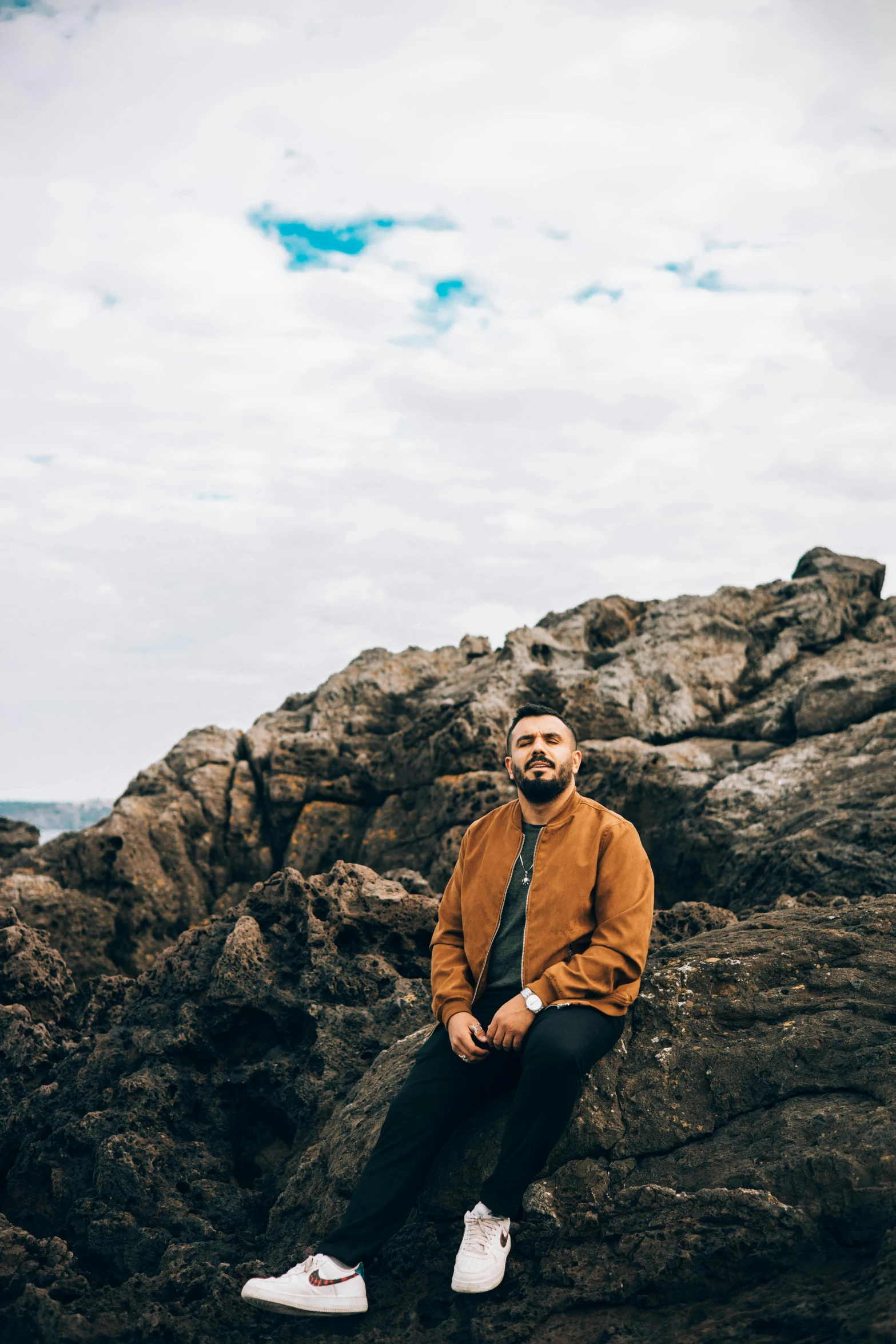 a man sitting on a rock near the ocean, an album cover, unsplash, portrait photo of a backdrop, burly man, he is wearing a brown sweater, graves