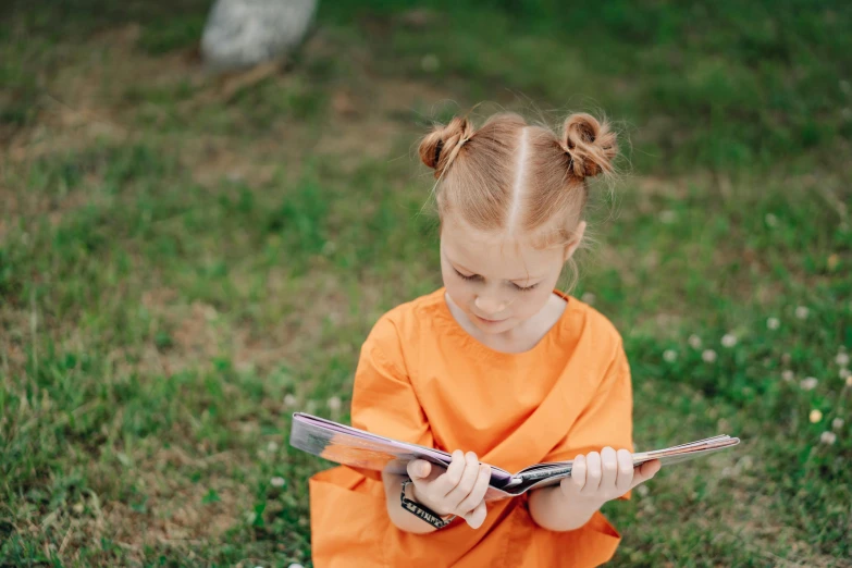 a little girl sitting in the grass reading a book, pexels contest winner, dressed in orange inmate attire, girl with messy bun hairstyle, gif, pictured from the shoulders up