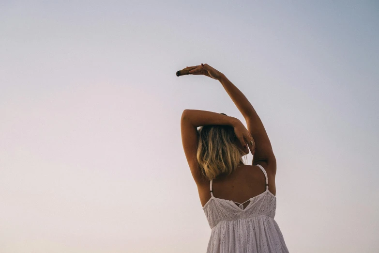 a woman in a white dress standing on a beach, unsplash, aestheticism, raising an arm, stretch, wearing a camisole, morning time