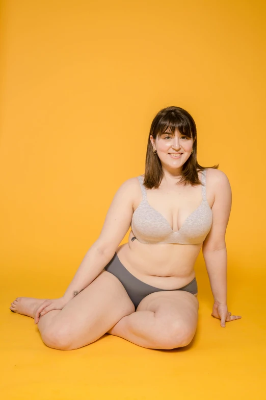 a woman in a bikini sitting on the floor, by Arabella Rankin, unsplash, yellow backdrop, she has a jiggly fat round belly, pale grey skin, posing together in bra