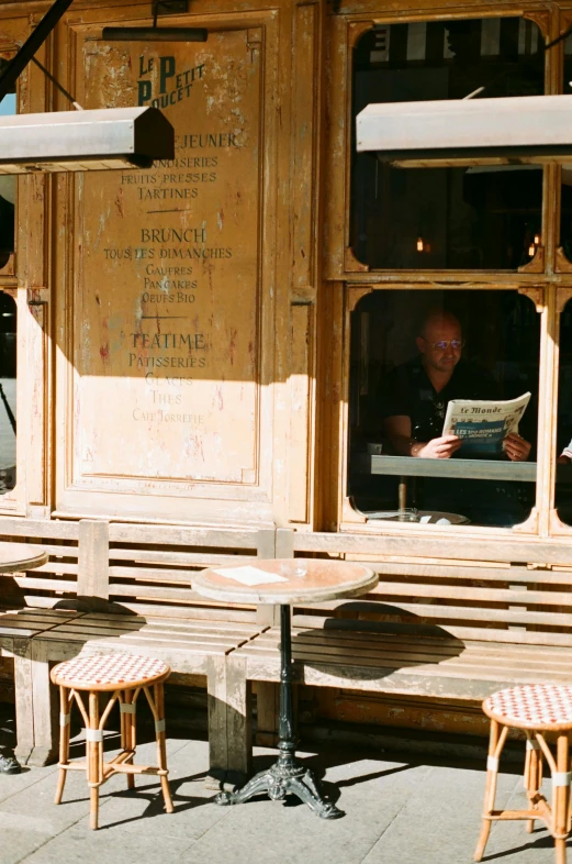 a man sitting at a table outside of a restaurant, a photo, by Jan Tengnagel, renaissance, sunny bay window, reading a newspaper, outside a saloon, wooden banks