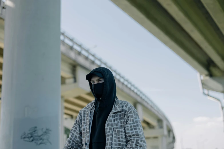 a man wearing a mask standing under a bridge, by Bascove, black hood, patterned clothing, filmstill, portrait featured on unsplash