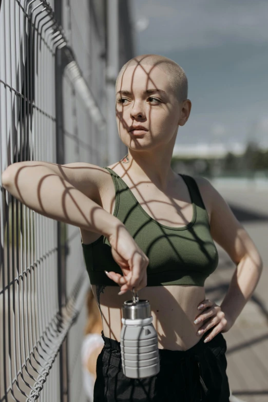 a woman with a shaved head leaning against a fence, by Adam Marczyński, pexels contest winner, detailed sports bra, holding a bottle, gif, action pose