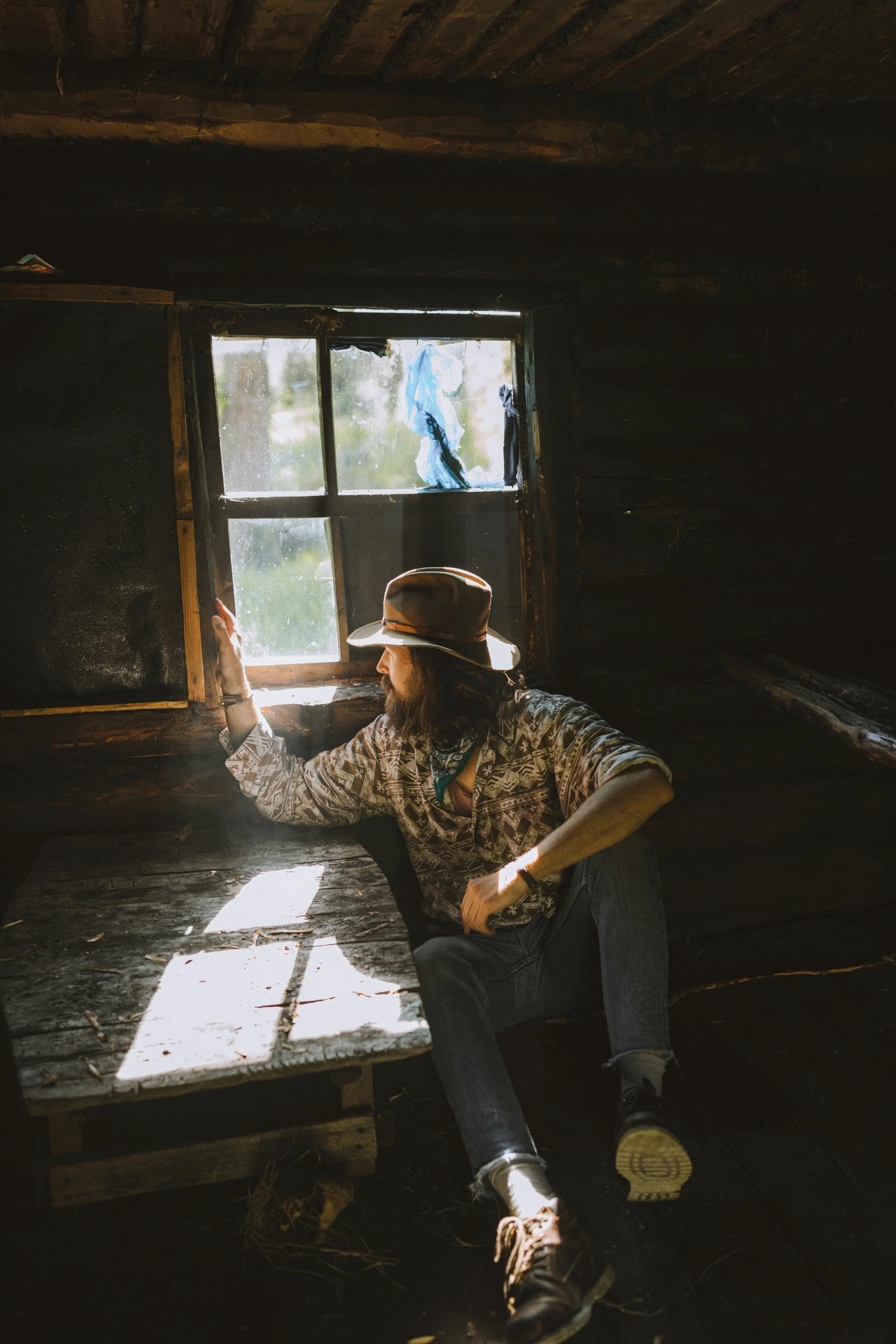 a man sitting on a bench in front of a window, by Jessie Algie, unsplash contest winner, australian tonalism, in an old west cabin, wears a destroyed hat, backlight glow, cottagecore hippie