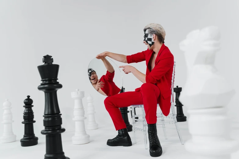 a man sitting on a chair next to a giant chess board, an album cover, by Emma Andijewska, pexels contest winner, red jumpsuit, xqc, looking into a mirror, zentai suit
