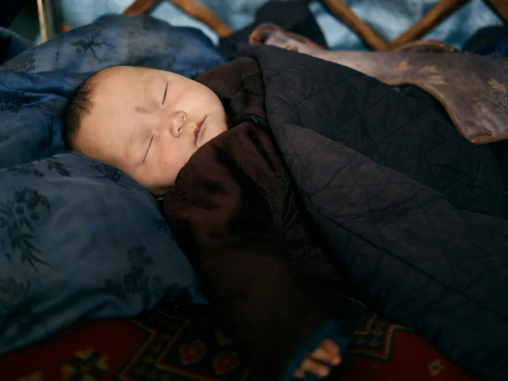 a baby sleeping in a blanket on top of a bed, by Nina Hamnett, pexels contest winner, shamanistic dark blue clothes, embroidered robes, model wears a puffer jacket, monk clothes