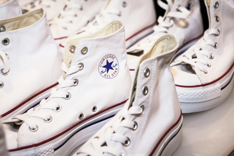 a row of white converse shoes sitting on top of a table, on display, high quality product image”