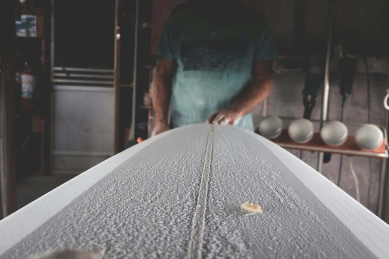 a man standing next to a surfboard in a garage, a stipple, unsplash, plasticien, long table, white wax, carving, thumbnail