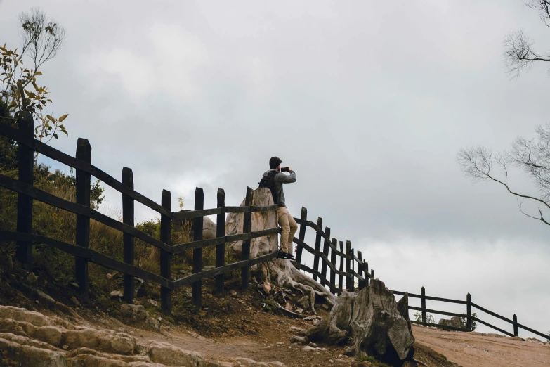 a man standing on top of a wooden fence, by Alejandro Obregón, unsplash, sitting on a rock, slight overcast weather, camera photo, multiple stories