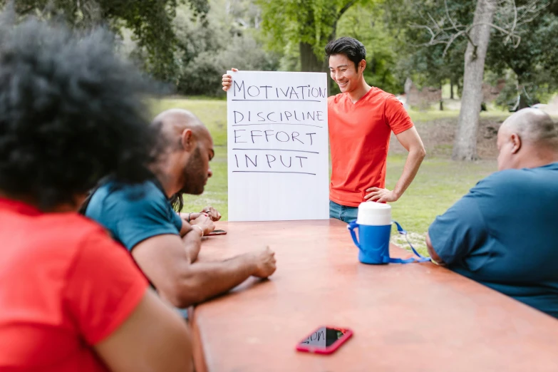 a group of people sitting around a table with a sign, pexels contest winner, at a park, motivation, a person standing in front of a, training