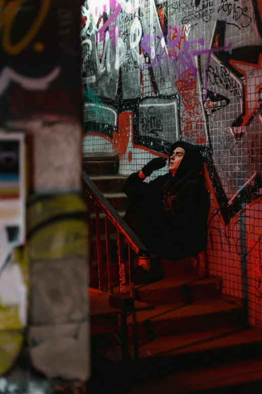 a person sitting on a set of stairs in front of a graffiti covered wall, inspired by Elsa Bleda, pexels contest winner, dressed in a black cloak, red mood in background, hyper - goth, resting