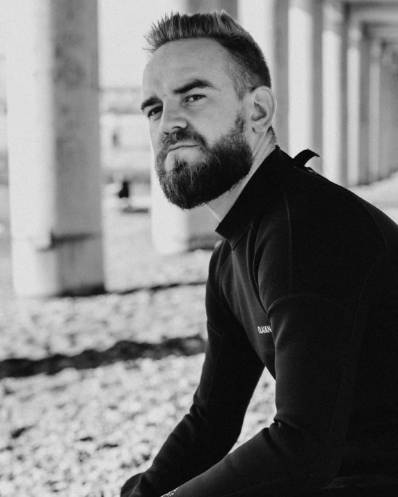 a man with a beard sitting on a bench, a black and white photo, inspired by Edi Rama, wearing black tight clothing, wearing a track suit, profile image, riding