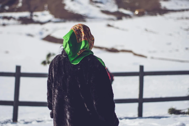 a person standing in front of a fence in the snow, trending on pexels, dressed in a green robe, wearing a head scarf, on the mountain, black