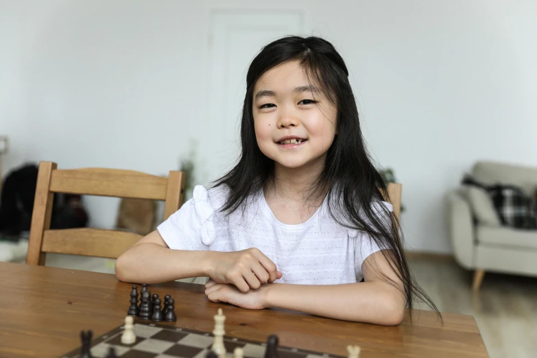 a little girl sitting at a table playing chess, a portrait, pexels contest winner, satisfied pose, taejune kim, smiling at camera, profile picture 1024px