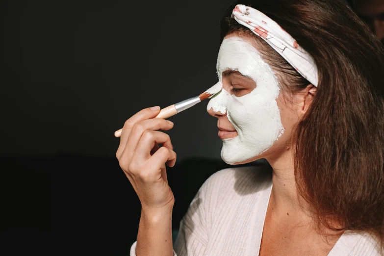 a woman putting a face mask on her face, by Julia Pishtar, natural make-up, white face paint, bottom angle, wearing a white robe