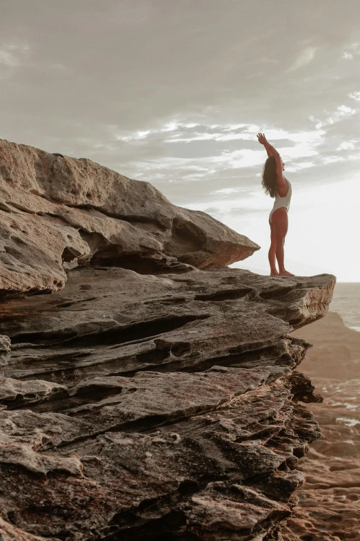 a woman standing on top of a cliff next to the ocean, by Jessie Algie, pexels contest winner, arabesque, in the australian outback, yoga pose, imbalanced mars. rugged, reaching for the sky