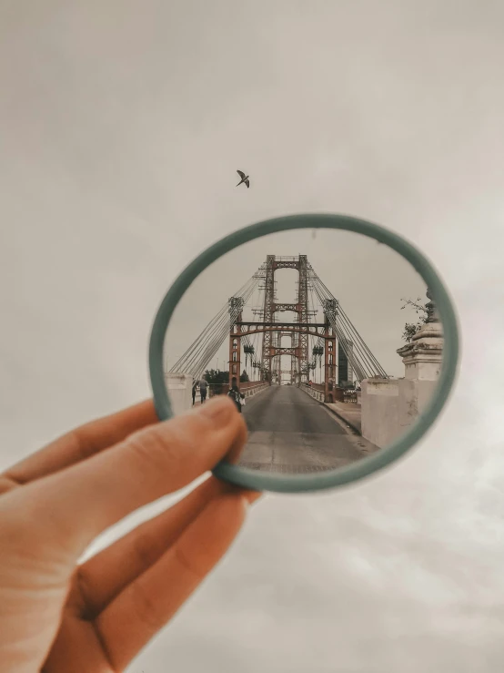 a person holding a magnifying lens with a bridge in the background, a picture, pexels contest winner, hyperrealism, instagram story, frame around picture, symmetrical shot, photo in color