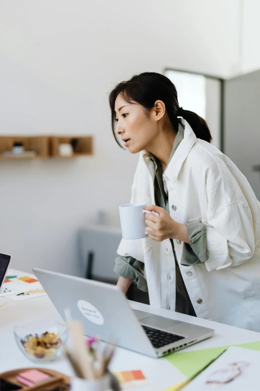 a woman sitting at a table with a laptop and a cup of coffee, by Jang Seung-eop, trending on pexels, happening, lab coat and tee shirt, looking confused, ethnicity : japanese, standing on a desk