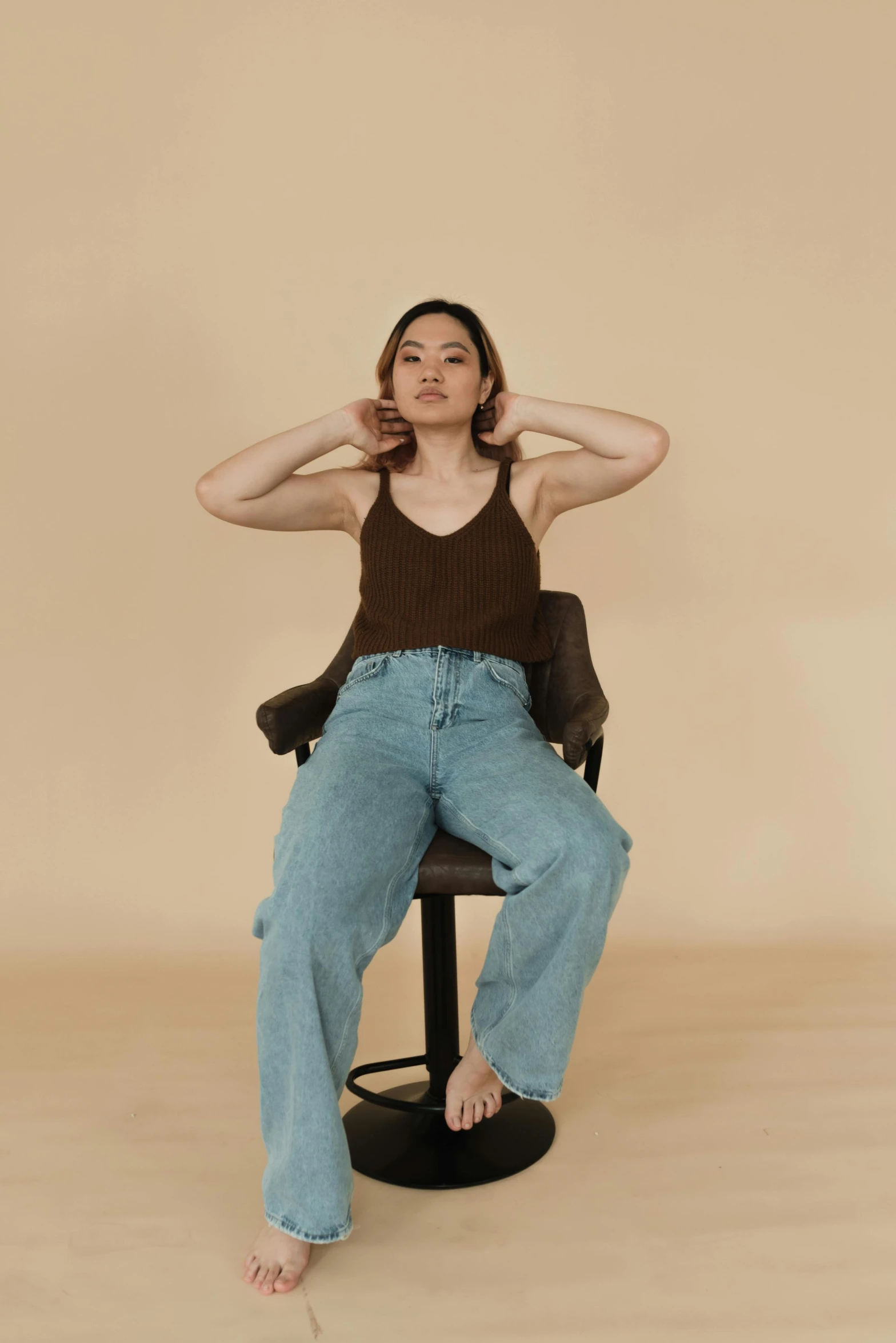 a woman sitting on a chair with her hands behind her head, by Robbie Trevino, trending on pexels, renaissance, baggy jeans, wearing : tanktop, wearing a brown, gemma chen