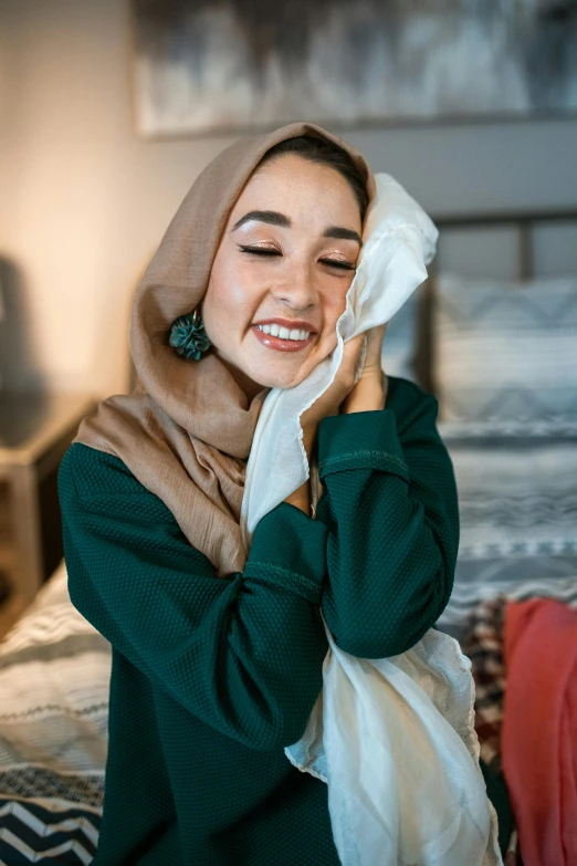 a woman sitting on a bed covering her face with a towel, by Julia Pishtar, hurufiyya, crying and smiling franticly, hijab, earing a shirt laughing, h3h3