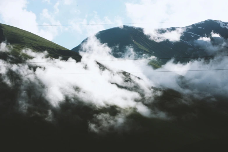 a train traveling through a lush green hillside, an album cover, pexels contest winner, hurufiyya, face made out of clouds, mountainous, overexposed photograph, black clouds