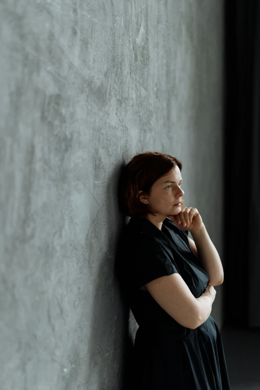 a woman in a black dress leaning against a wall, inspired by Anna Füssli, worried, grey backdrop, in a room, alison hannigan