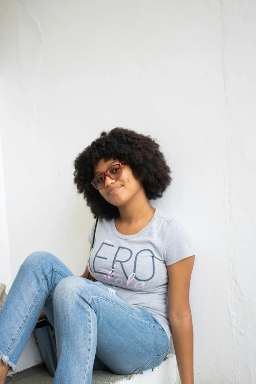 a woman sitting on top of a white stool, by Leo Michelson, pexels contest winner, afrofuturism, wearing a t-shirt, cute slightly nerdy smile, fair curly hair, grey