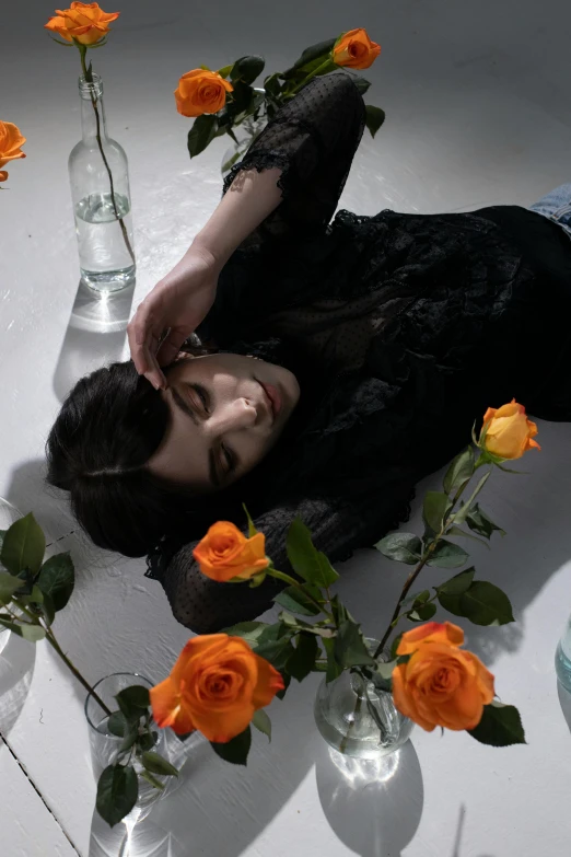 a woman laying on the floor surrounded by orange roses, androgynous person, goth aesthetic, with black hair, on a white table