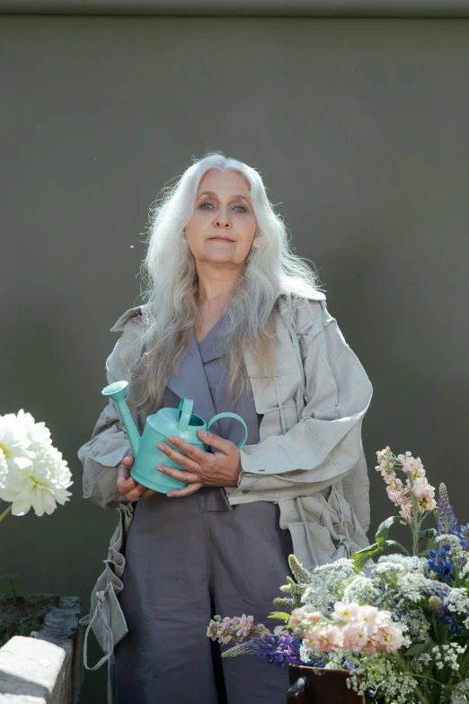 a woman standing next to a bunch of flowers, inspired by Grethe Jürgens, light gray long hair, watering can, catalogue photo, the look of an elderly person