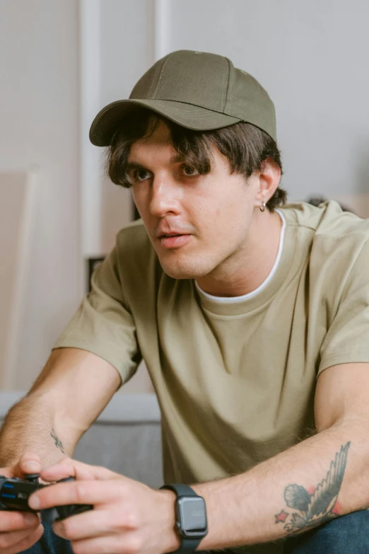 a man sitting on a couch holding a game controller, a colorized photo, inspired by John Luke, in a dark green polo shirt, symmetrical face orelsan, wearing a backwards baseball cap, sitting on a mocha-colored table