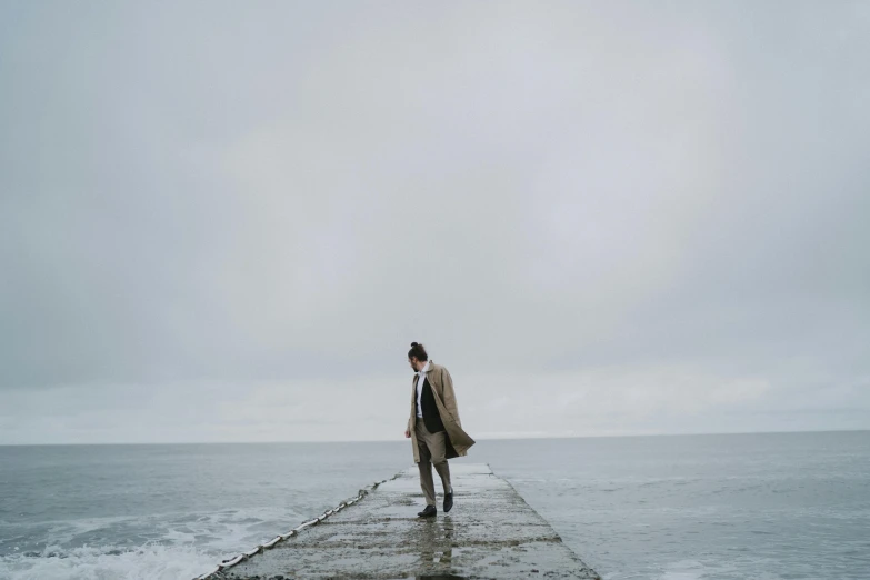 a man standing on a pier next to the ocean, a picture, unsplash, trench coat and suit, gray sky, ignant, a woman