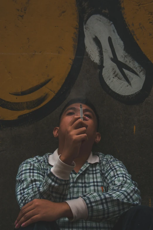 a man sitting in front of a wall with a smiley face painted on it, a picture, pexels contest winner, cigarette dangling, teenage boy, forward facing, slight haze