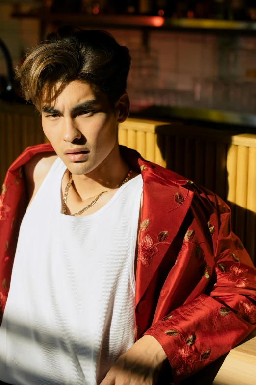 a man in a red jacket sitting at a table, an album cover, inspired by John Luke, trending on pexels, asian man, at a fashion shoot, jewelry, late afternoon