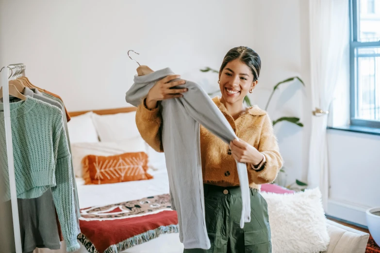 a woman standing in front of a bed holding a shirt, pexels contest winner, happening, pokimane, inspect in inventory image, smiling, wearing a grey robe