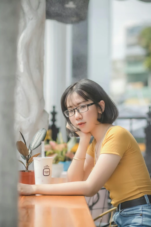 a woman sitting at a table with a cup of coffee, by Tan Ting-pho, pexels contest winner, realism, wearing yellow croptop, wearing square glasses, young adorable korean face, thoughtful )