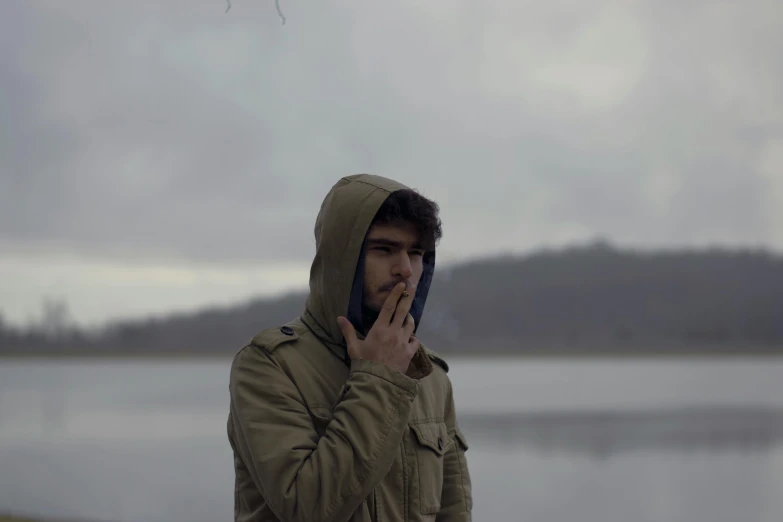 a man standing in front of a lake talking on a cell phone, by Attila Meszlenyi, pexels contest winner, hyperrealism, cigarette in his mouth, hooded, movie still of a tired, zayn malik