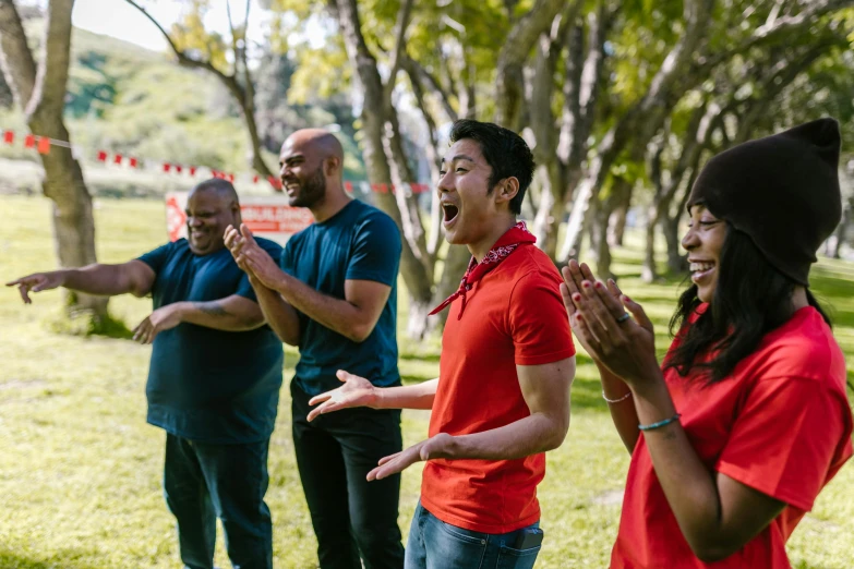 a group of people standing on top of a lush green field, throwing cards in the air, wearing a red hoodie, varying ethnicities, sing for the laughter