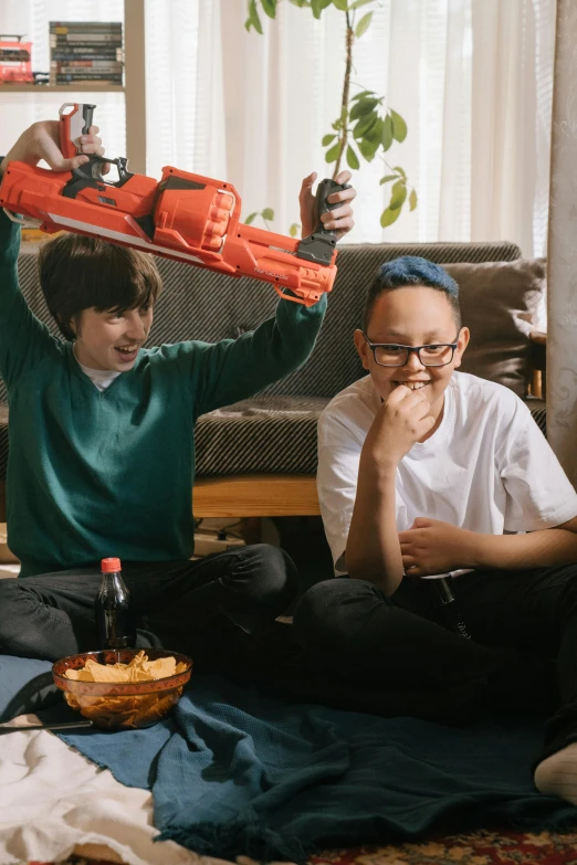 a group of people sitting on top of a living room floor, next to it is a toy ray gun, mukbang, brunette boy and redhead boy, promo image